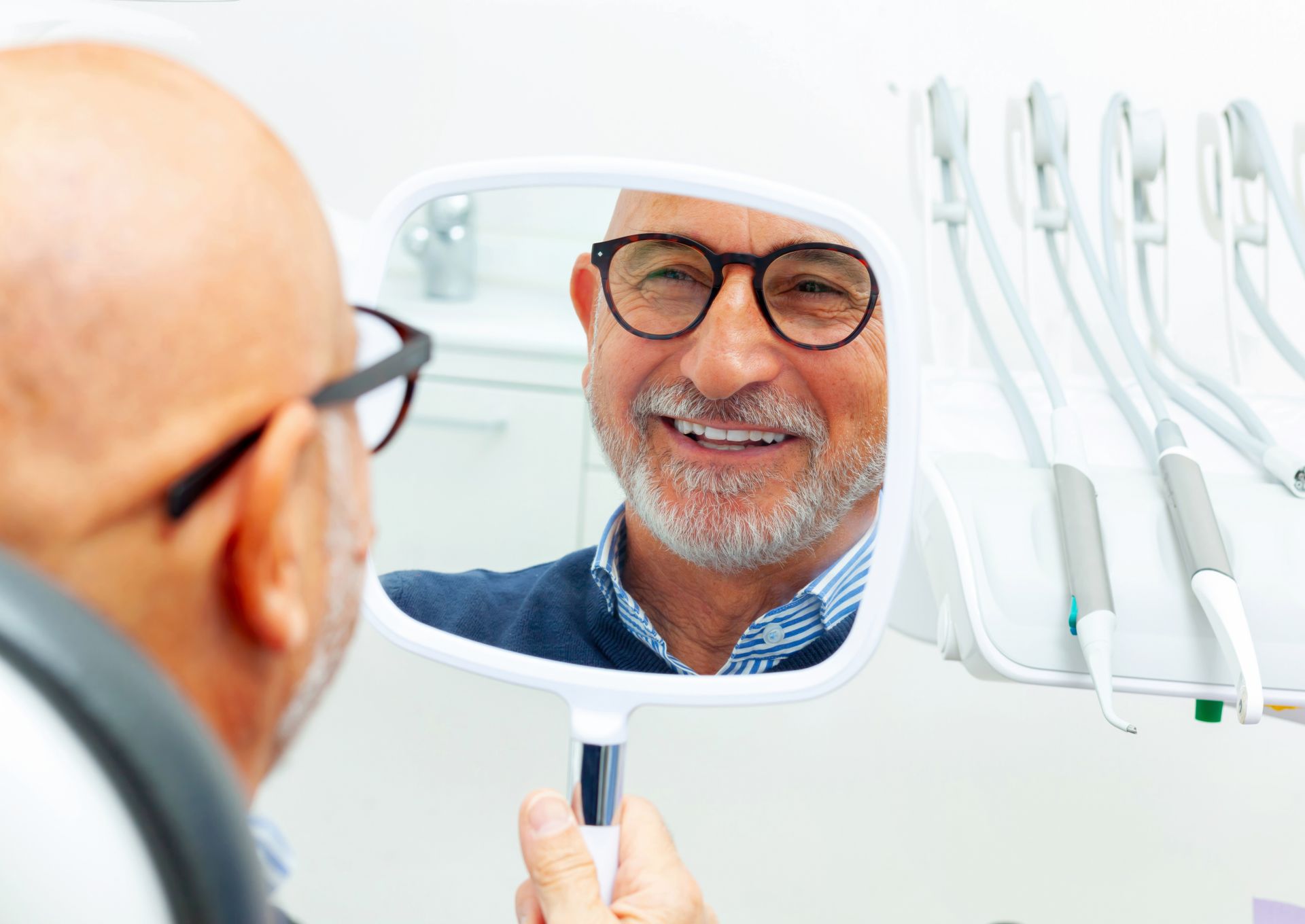 Man in glasses smiling, looking at his teeth in a handheld mirror in a dental office.