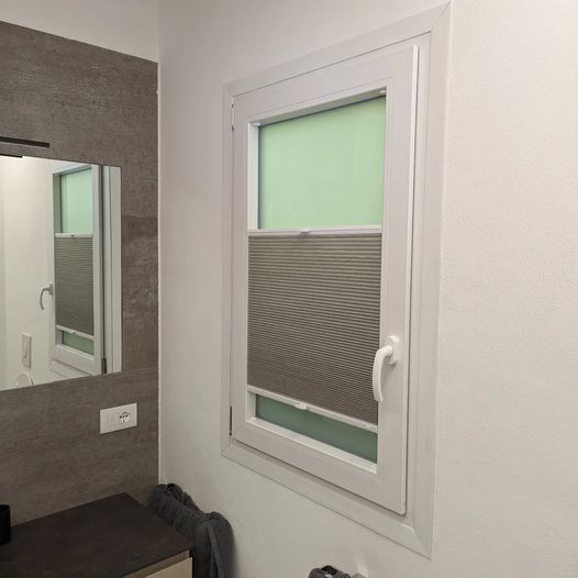 Finestra bianca con tende plissettate parzialmente abbassate in un bagno.