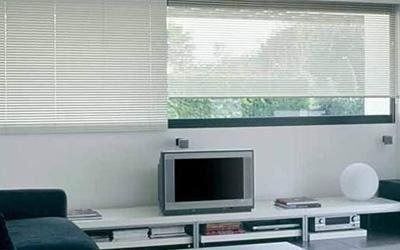 Soggiorno con TV, tende orizzontali, parete bianca e scaffali.