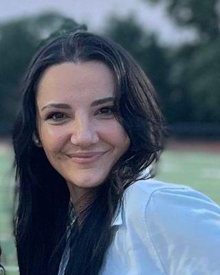Woman with dark hair smiles, wearing a white shirt; set outdoors with a blurred green field in background.