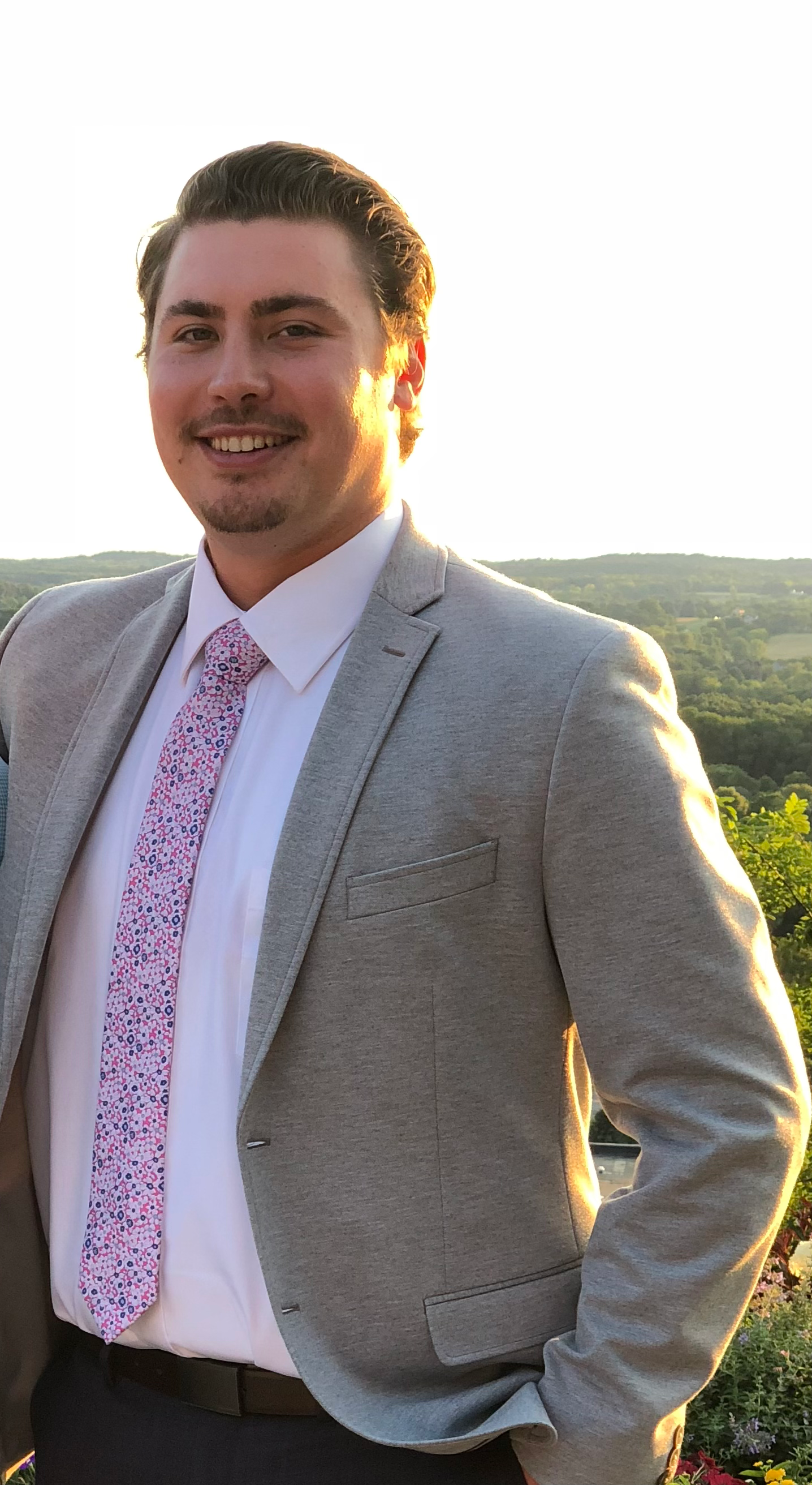 Man in grey blazer and floral tie smiles outdoors.