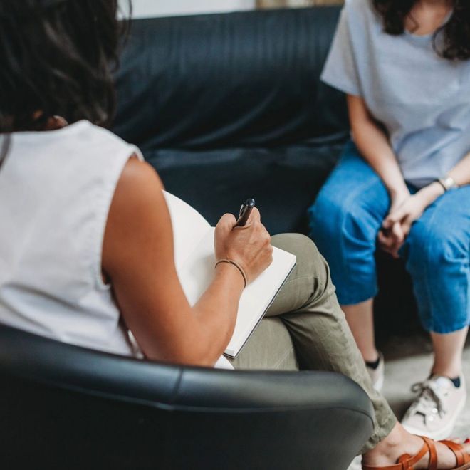Therapist taking notes as client sits on couch. Indoors, focus on their conversation.