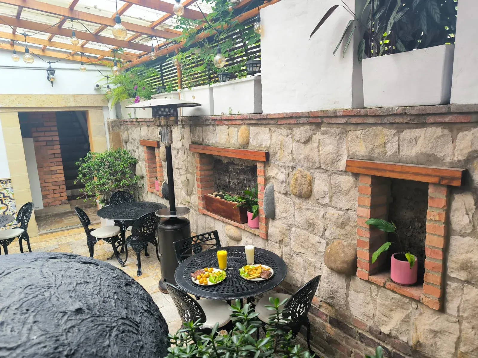 Comedor al aire libre con muro de piedra, mesas, sillas, plantas y calefactor.