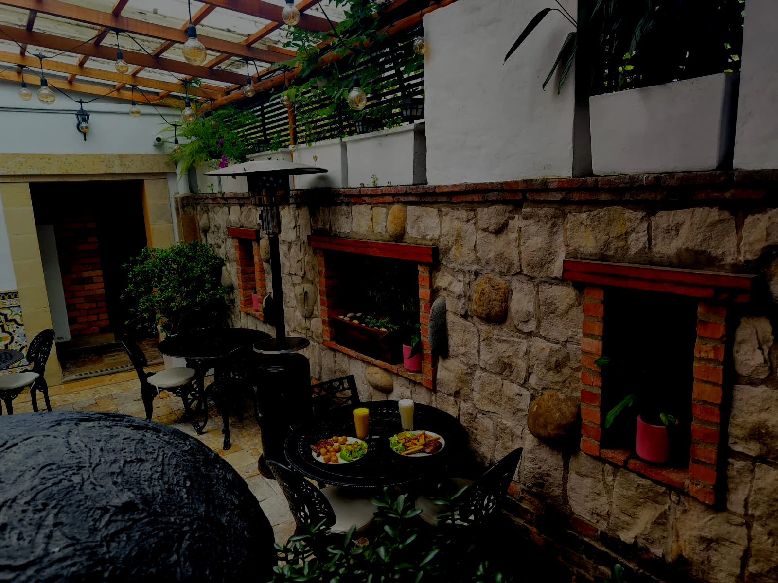 Outdoor cafe seating area with stone wall, wooden pergola, and food on a table.