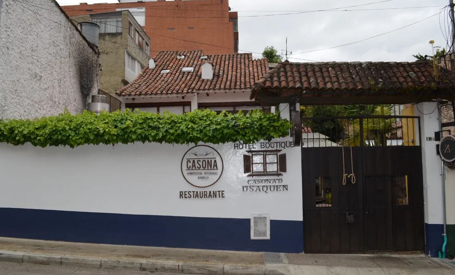 Fachada del restaurante con pared blanca y azul, puerta de madera y toldo cubierto de enredaderas.