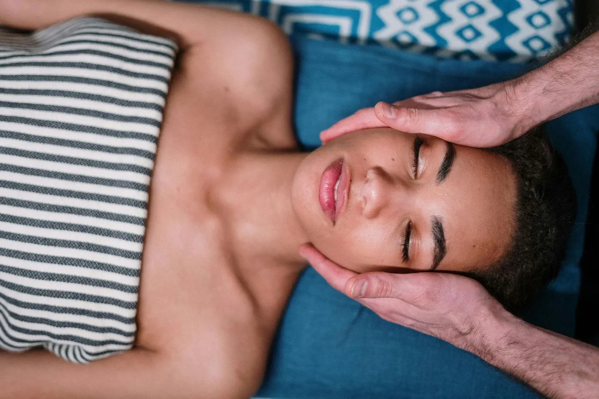 Mujer recibiendo masaje facial, acostada sobre una almohada azul, envuelta en una toalla a rayas.