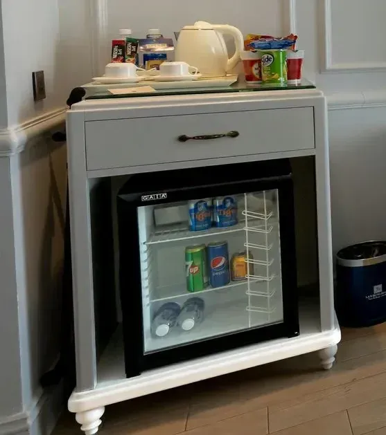 Pequeño armario de hotel con minibar que contiene bebidas y estante con té y aperitivos.