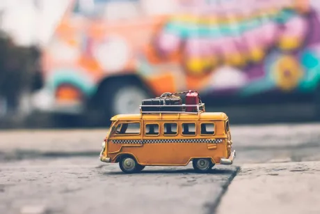 Autobús de juguete amarillo con equipaje en el techo, furgoneta colorida borrosa al fondo, en una calle.