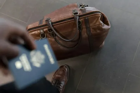 Persona sosteniendo un pasaporte cerca de una bolsa de viaje de cuero marrón y un zapato marrón sobre un suelo oscuro.