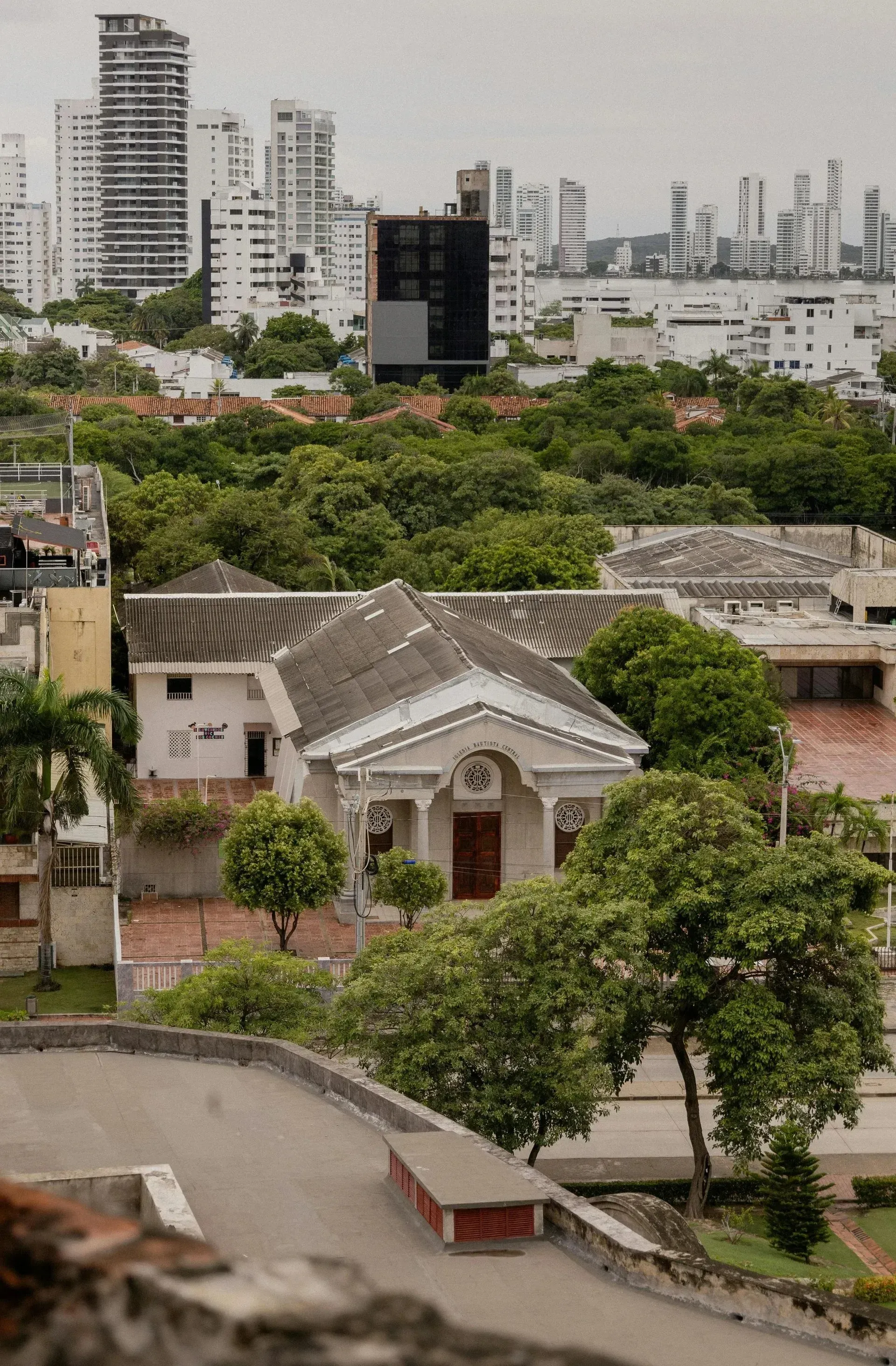 Vista de paisaje urbano con un edificio colonial blanco, rodeado de árboles verdes, con rascacielos a lo lejos.
