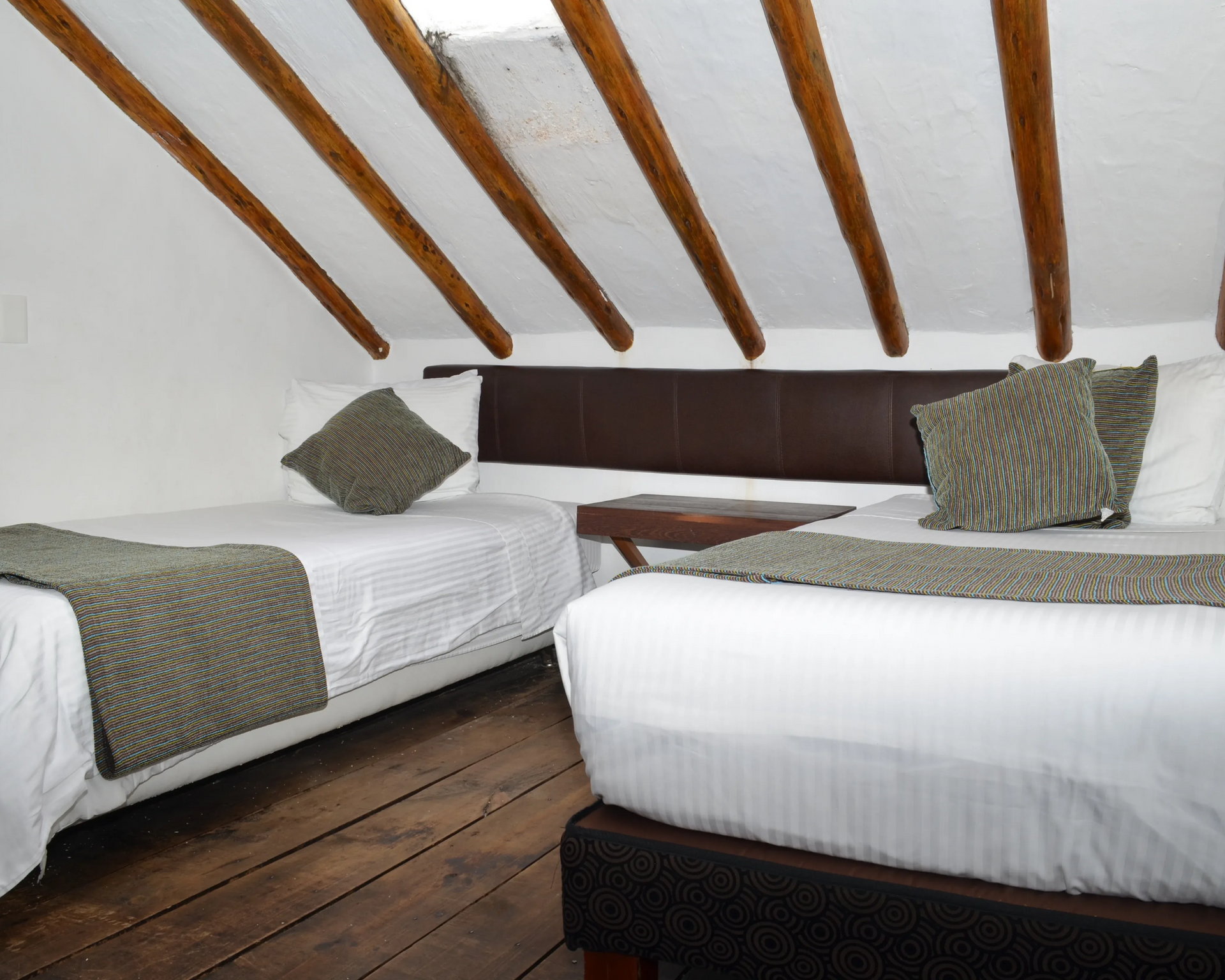 Dormitorio con dos camas bajo un techo inclinado con vigas de madera, ropa de cama blanca y cabecero marrón.