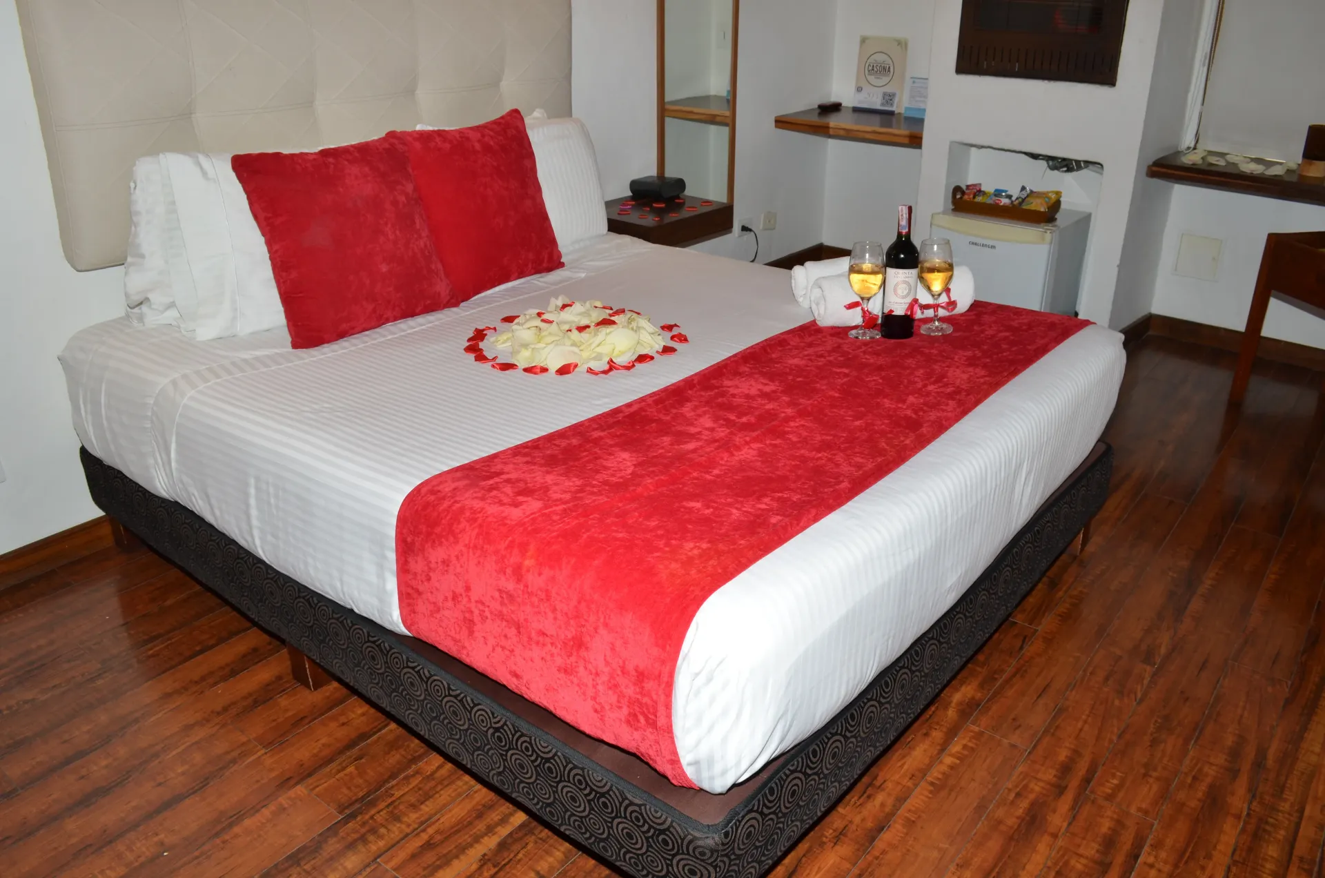 Habitación de hotel con cama. Alfombra y almohadas rojas, pétalos de rosa, vino y aperitivos sobre la cama.