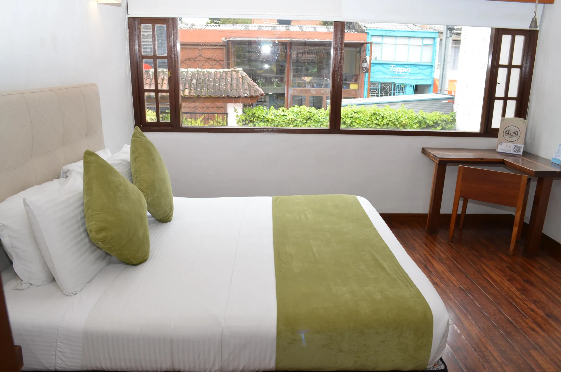 Dormitorio con cama blanca, detalles verdes, ventana con vistas a la calle y escritorio de madera.