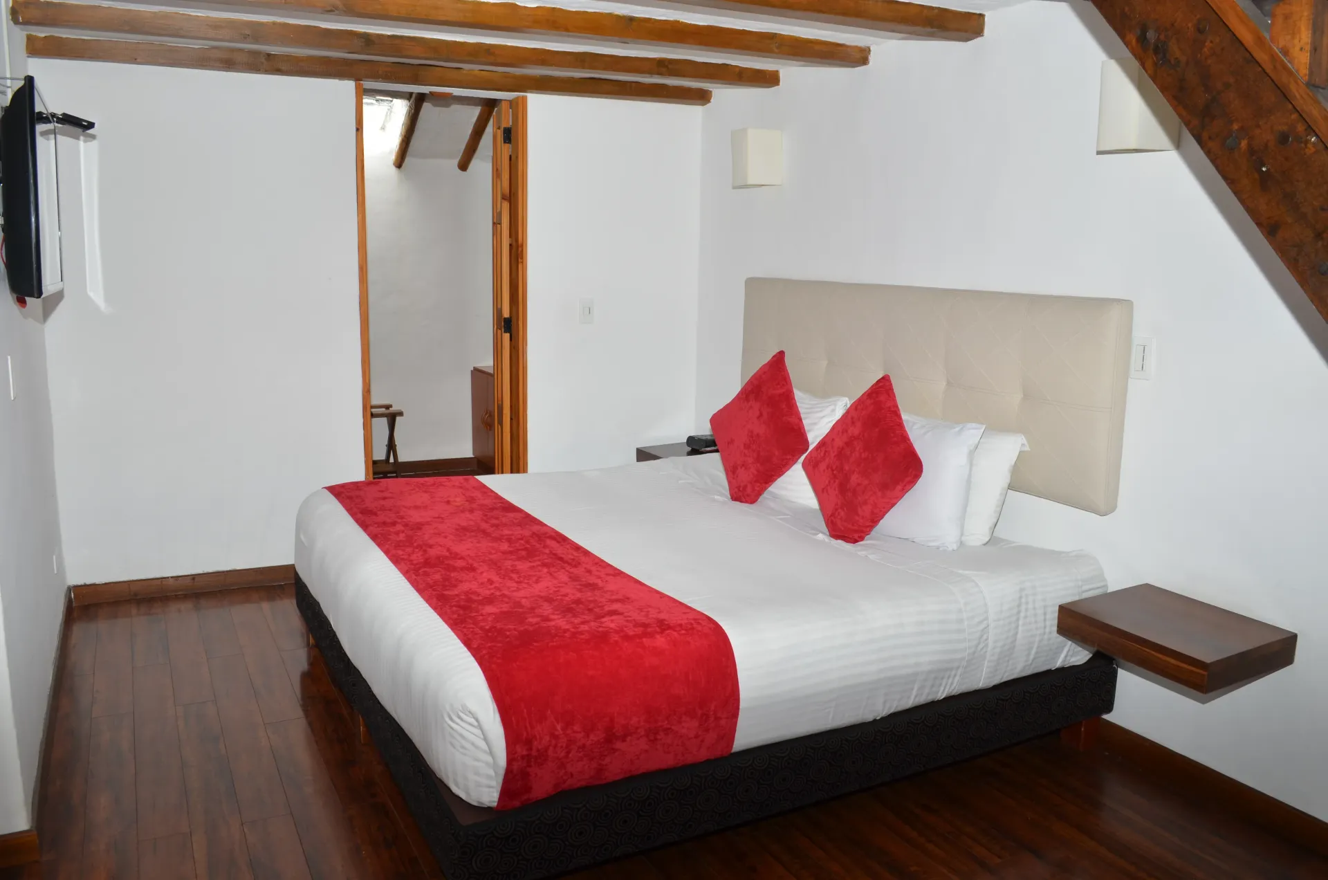 Dormitorio con cama, detalles en rojo y vigas de madera. Puerta abierta al baño.