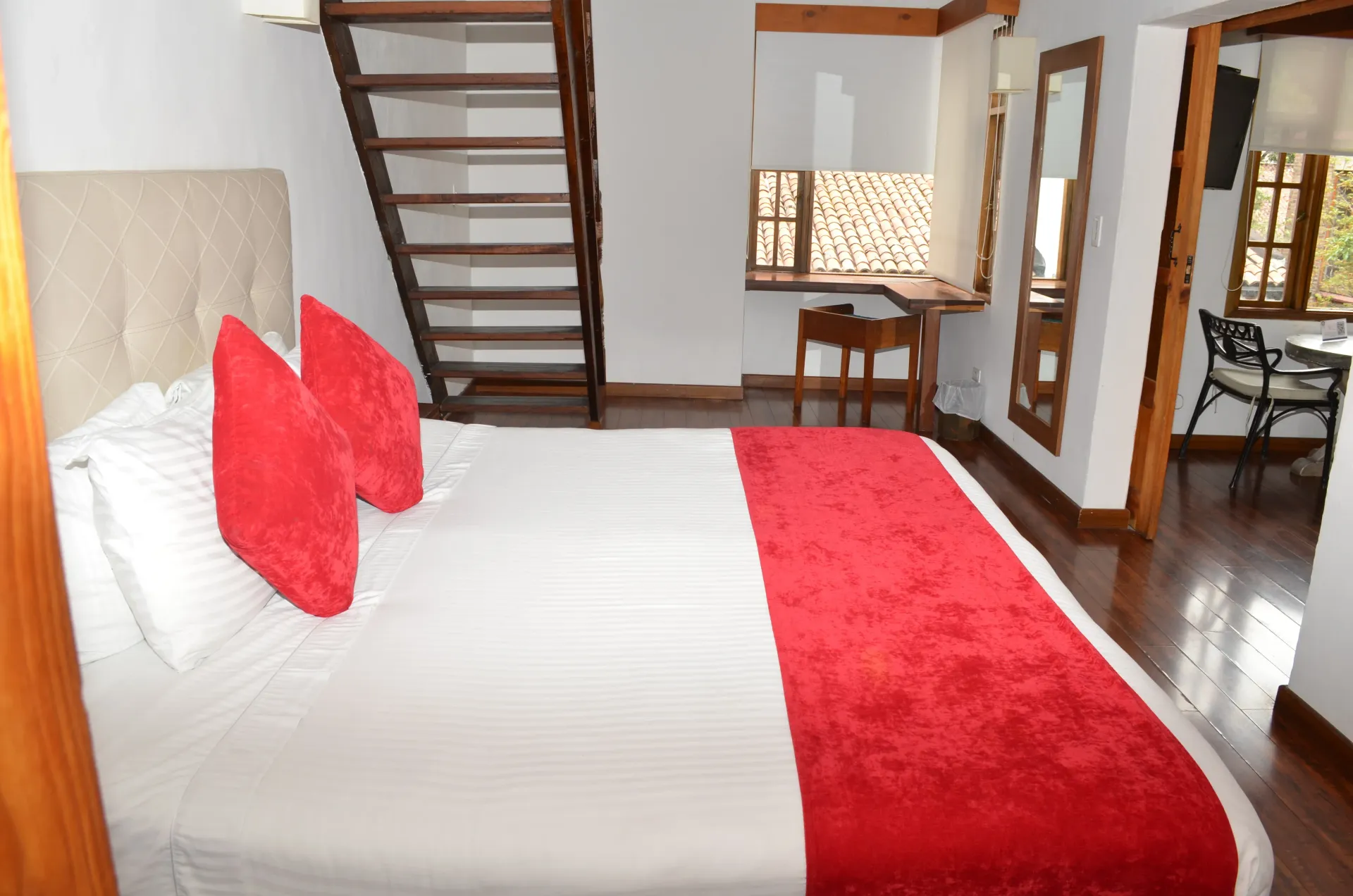 Dormitorio con cama, almohadas rojas y alfombra, escalera de madera, escritorio y puerta.