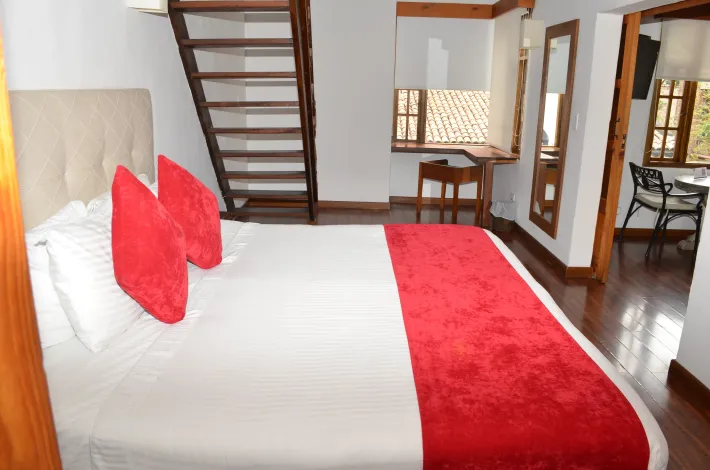 Dormitorio con cama, almohadas rojas y alfombra, escalera de madera y un pequeño escritorio.