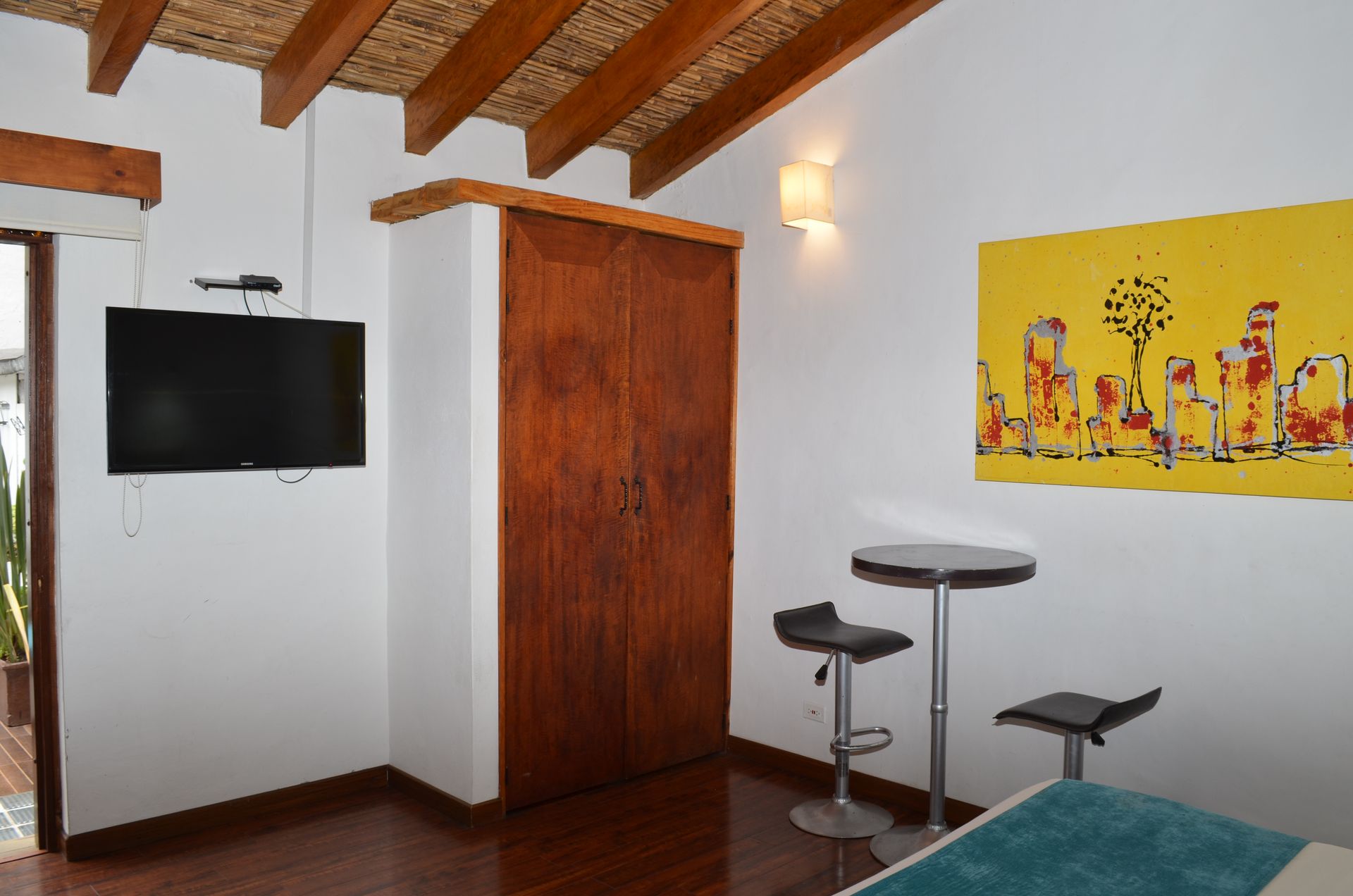 Habitación con televisor, armario de madera, mesita con taburetes y cuadro de un paisaje urbano amarillo.