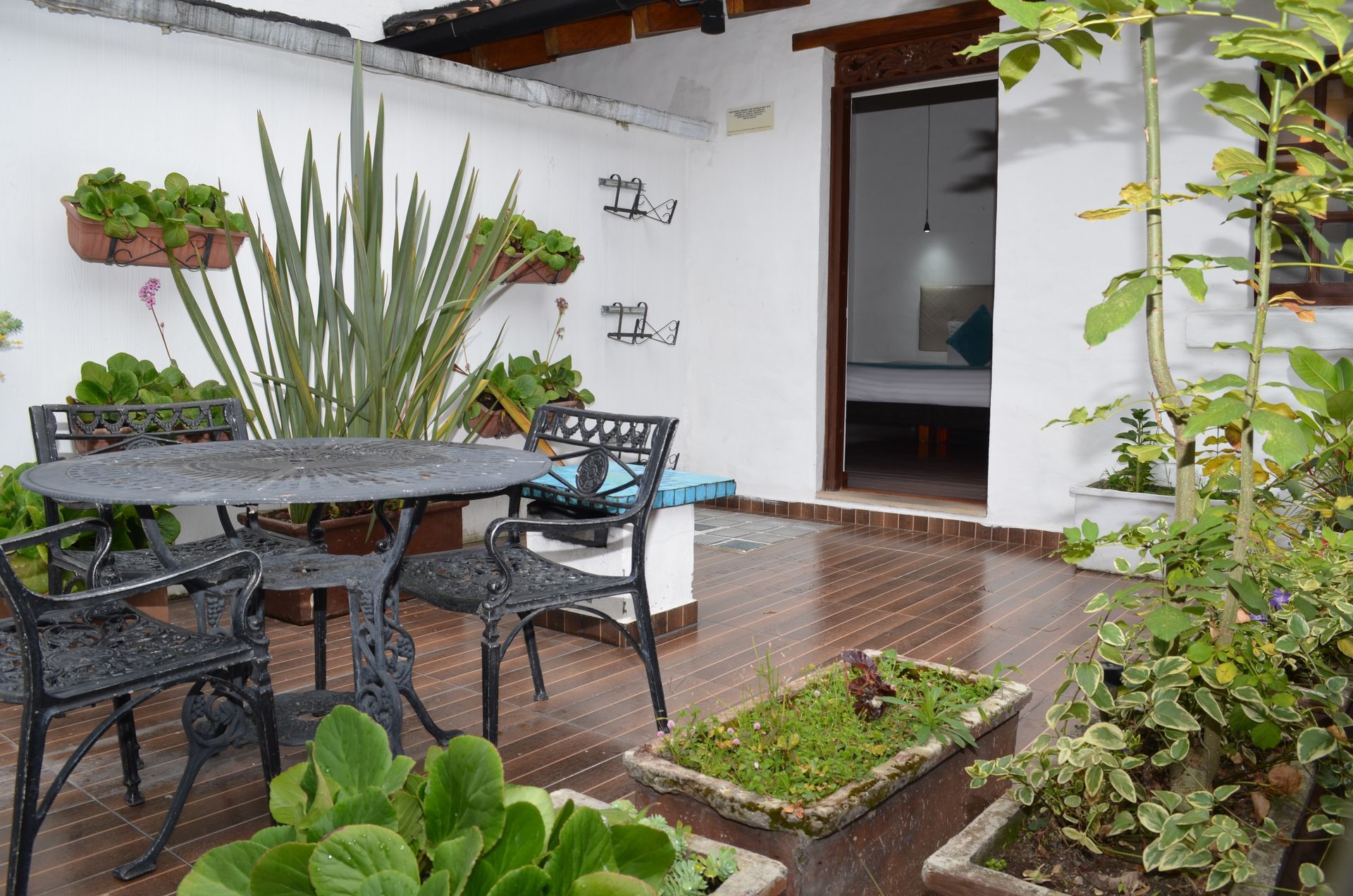 Patio con mesa y sillas de metal negro, plantas en macetas y puerta de acceso a un dormitorio.