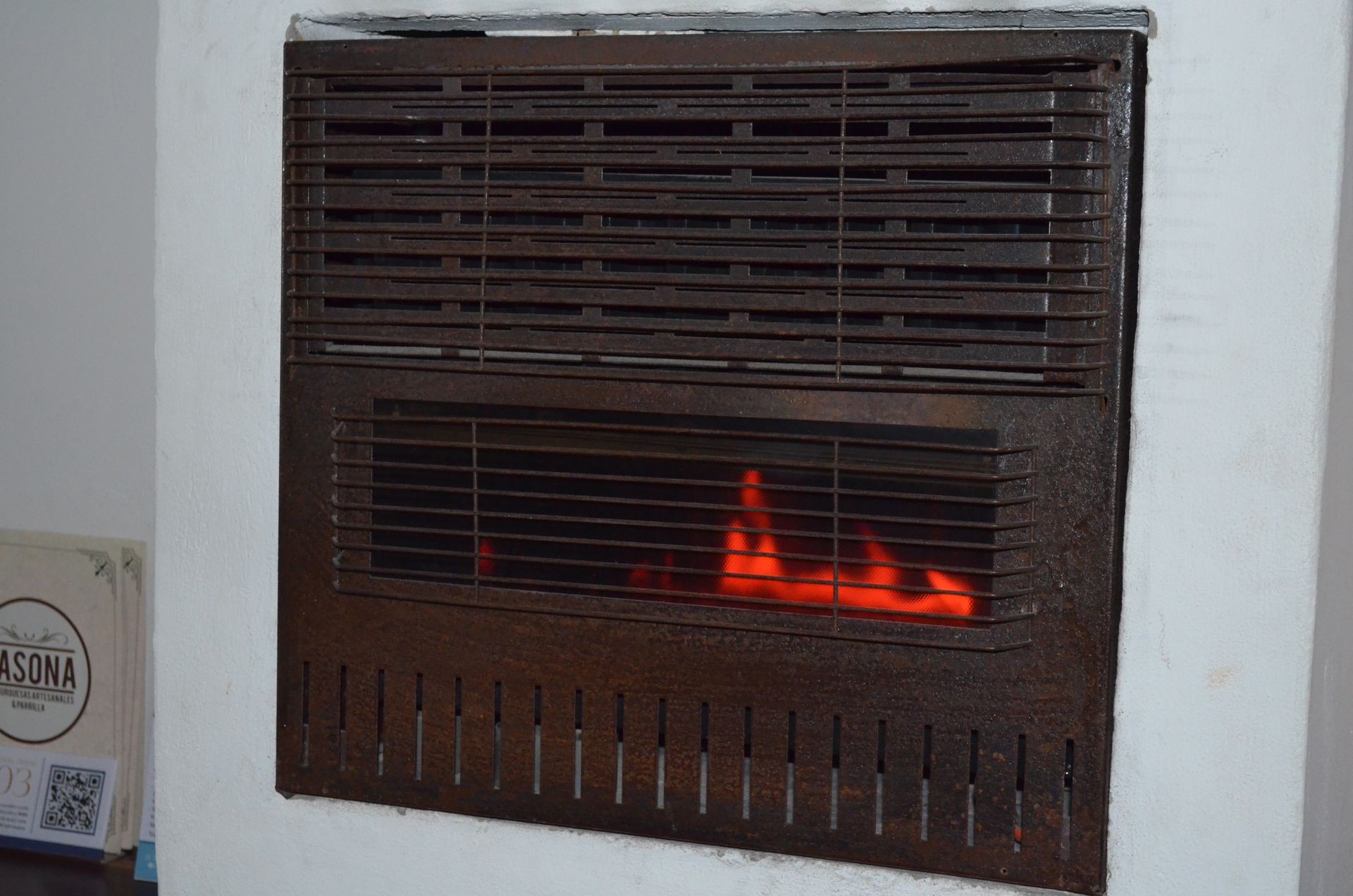 Un calentador de gas mural con una llama naranja visible, empotrado en una pared blanca.