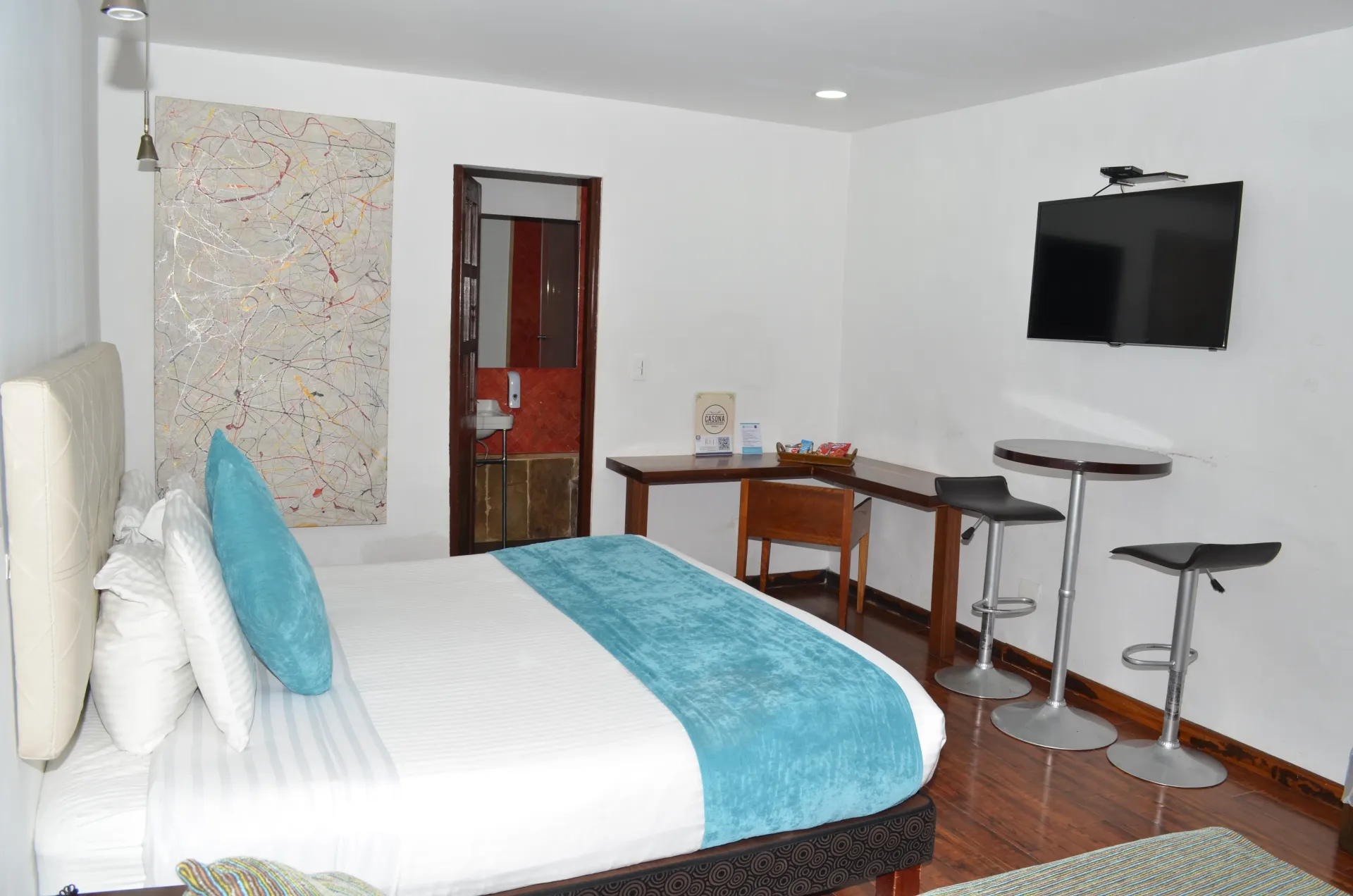 Habitación de hotel con cama, escritorio, taburetes de bar y televisor. Paredes blancas, colcha azul y suelo de madera.