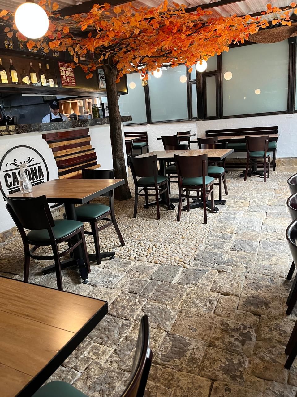Terraza del restaurante con mesas, sillas, árbol con hojas naranjas, suelo de piedra y grandes ventanales.