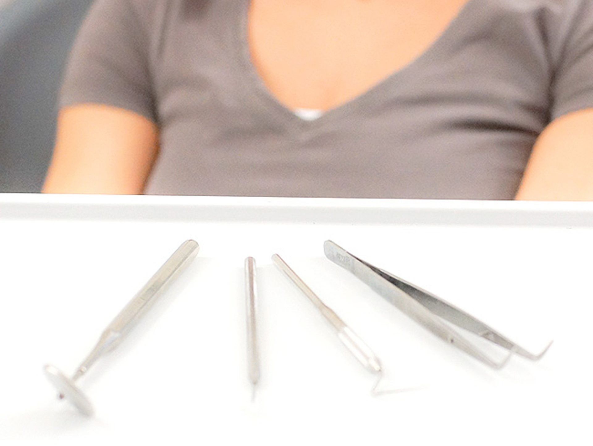 A woman in a grey shirt sits in front of a tray of tweezers