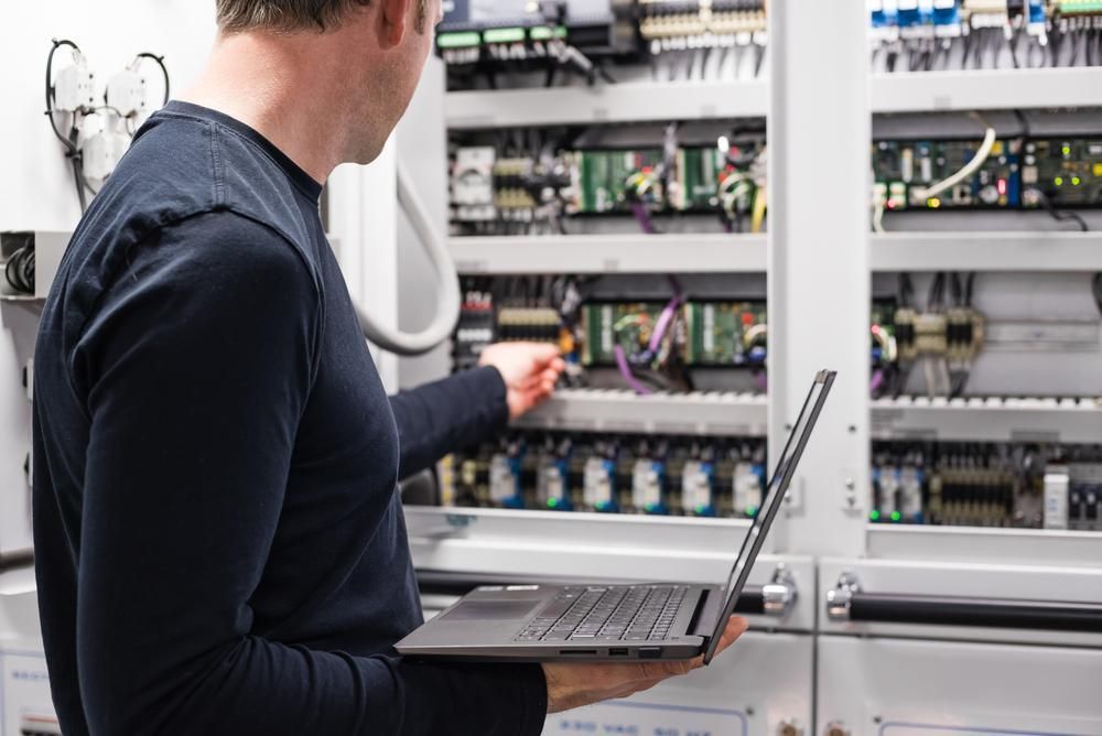A Man is Holding a Laptop in Front of a Electrical Panel — Power & Data Support Services In Karumba, QLD