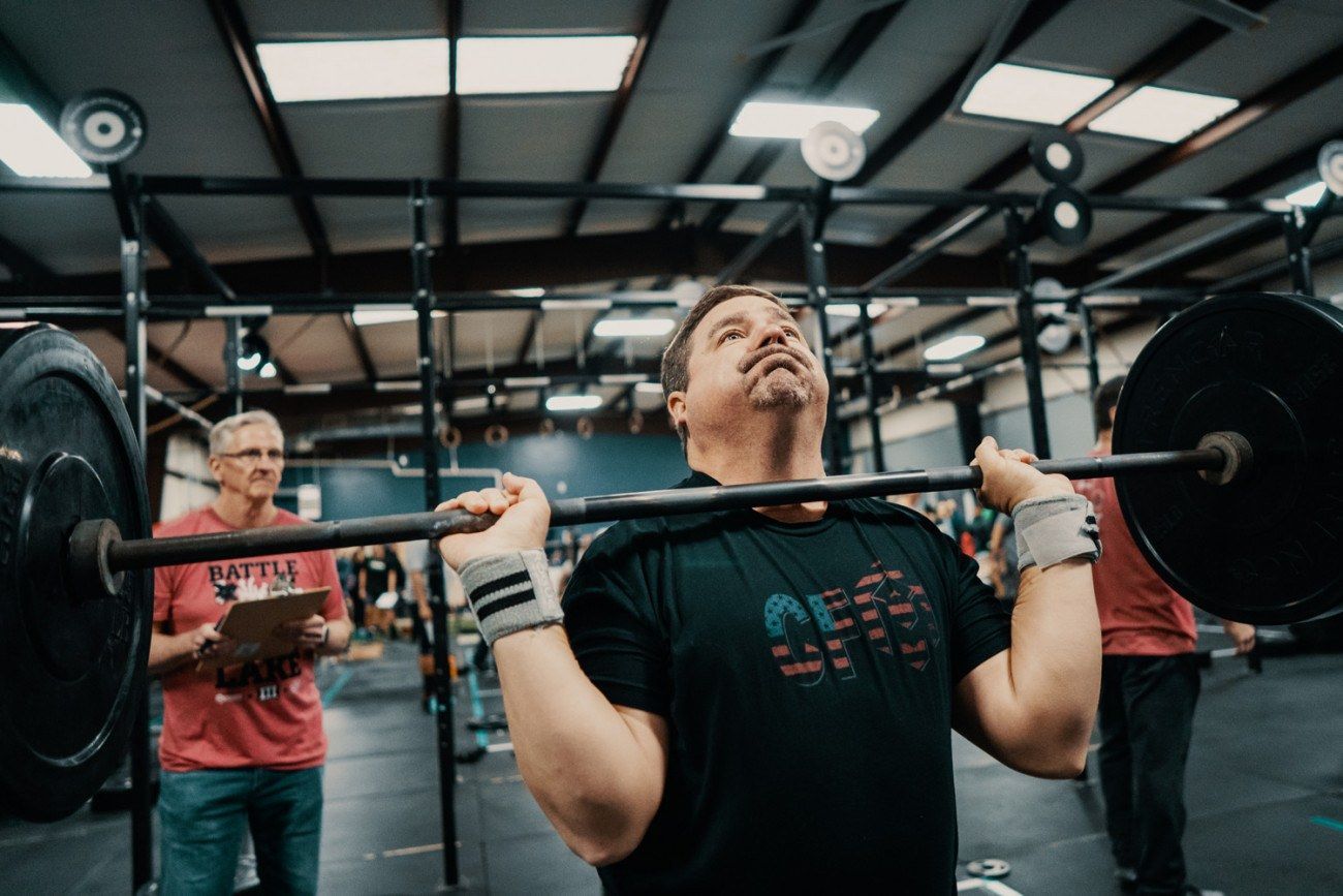 Man lifting barbell overhead in gym, focused expression. Coach watches.