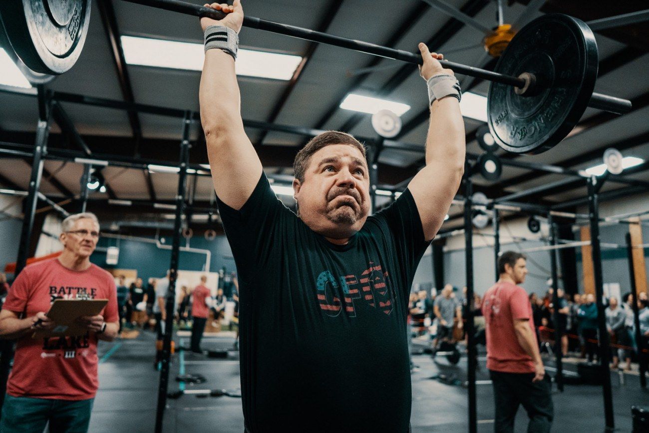 Man lifting barbell overhead in a gym; others watching in the background.