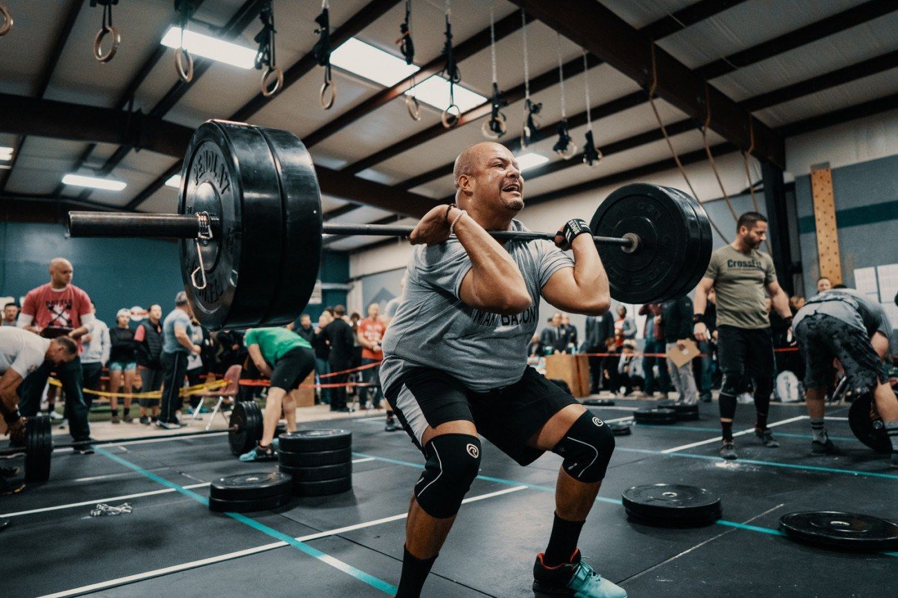 Weightlifter in gray shirt performs a clean and jerk, lifting a heavy barbell in a gym.
