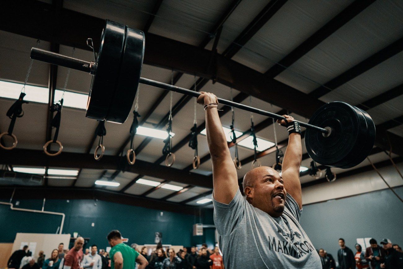 Man lifting heavy barbell overhead in a gym, surrounded by people, with a look of exertion and joy.