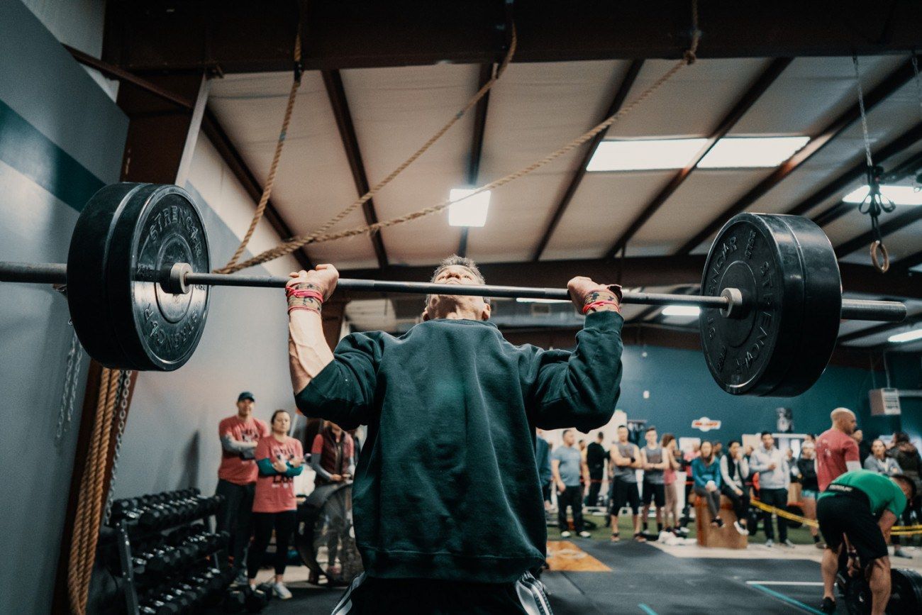 Person lifting a barbell overhead in a gym, with onlookers.