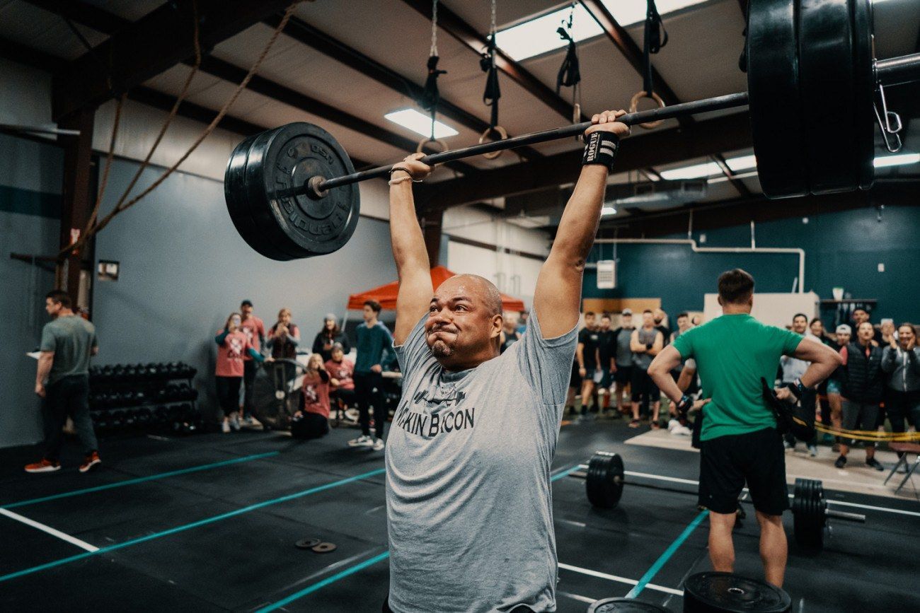 Man in gray shirt lifts a heavy barbell overhead in a gym, with a crowd watching.
