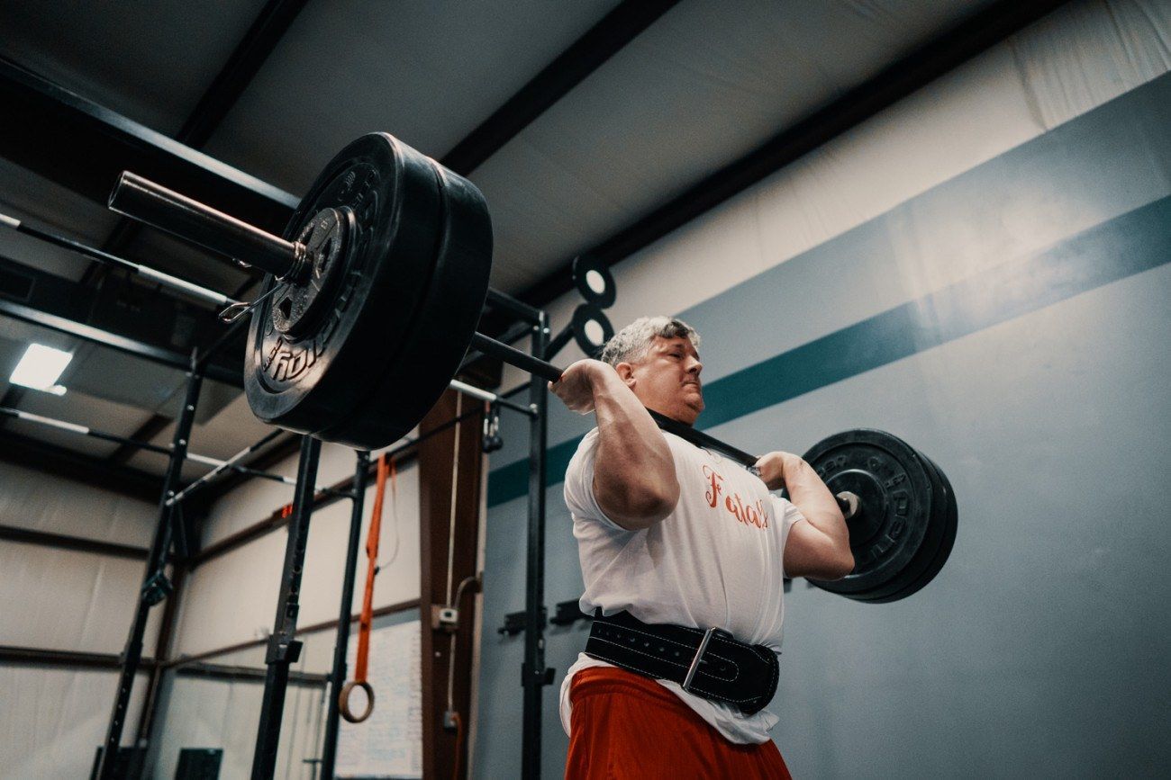 Man lifting a heavy barbell in a gym. He wears a weight belt and red shorts.