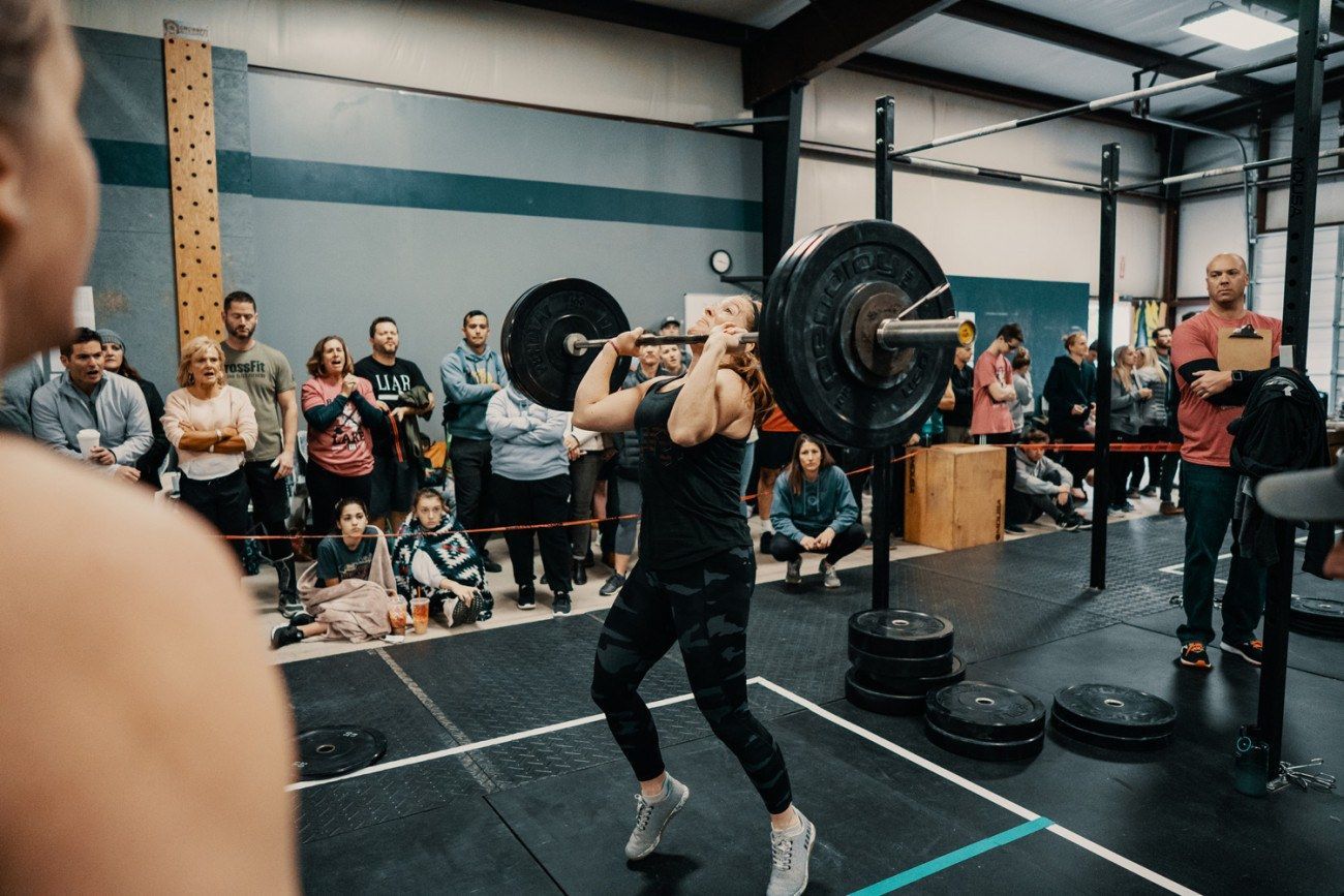 Woman in black lifting barbell overhead in a gym competition; crowd watches.
