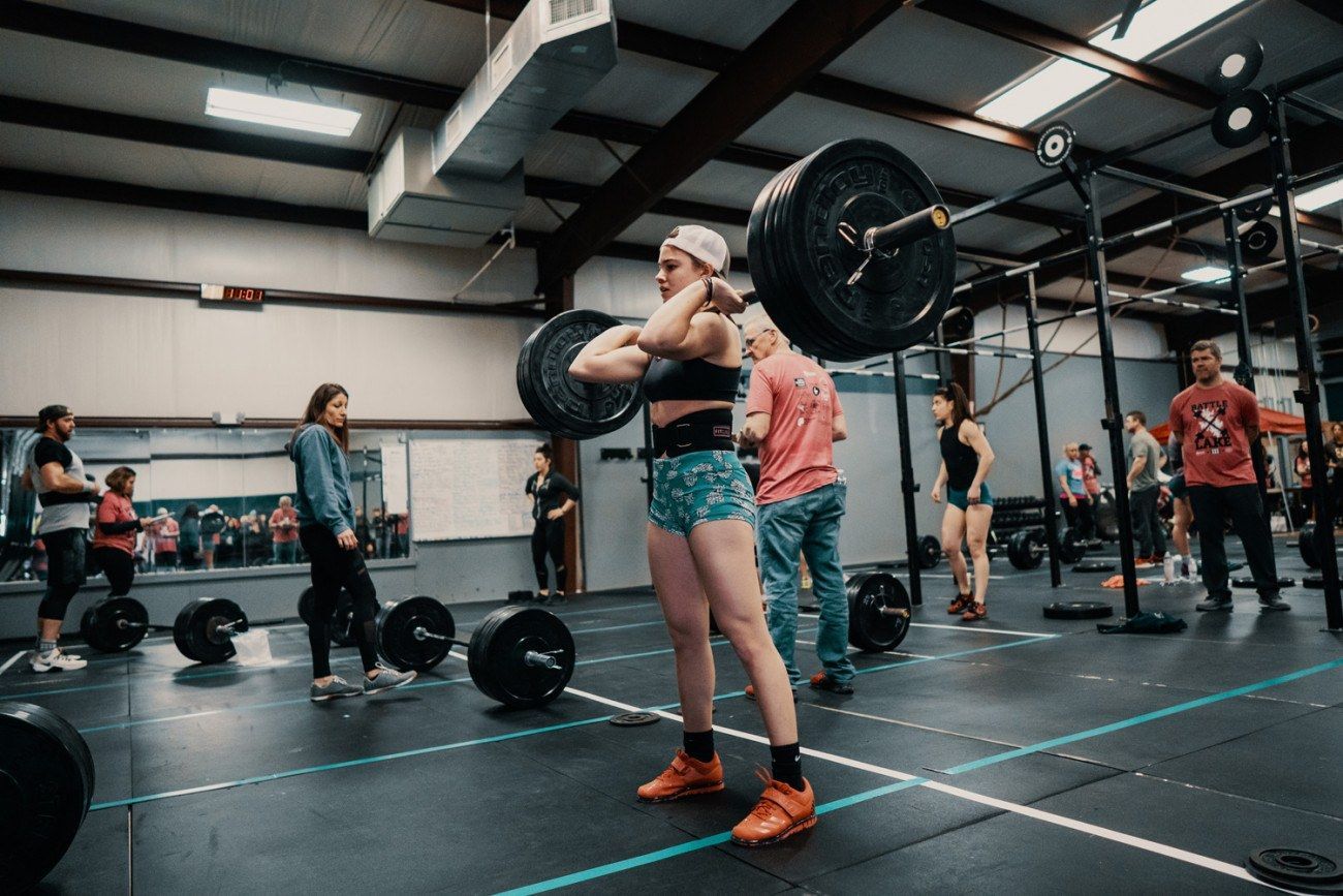 Woman in gym with barbell, hands crossed, facing forward. Others nearby.