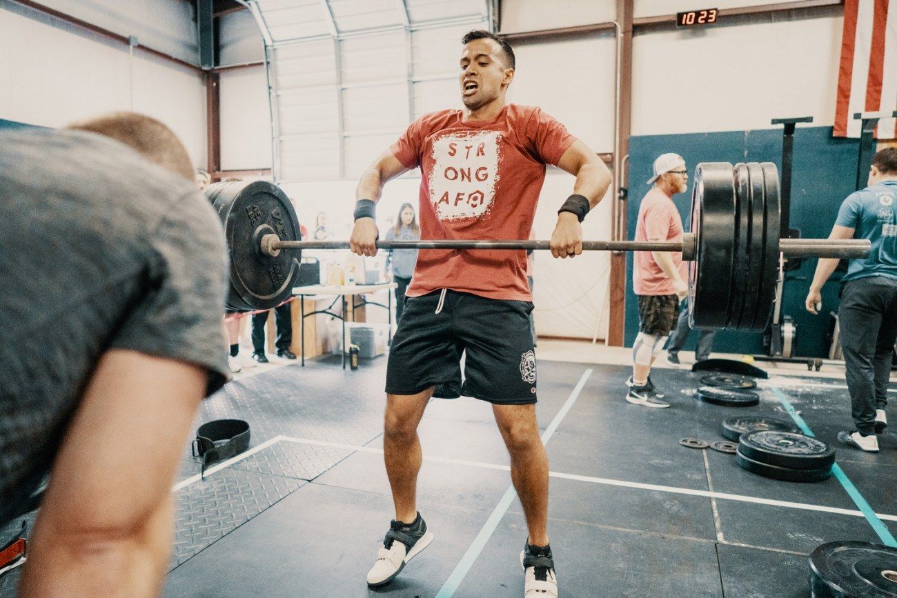 Man lifting a heavy barbell in a gym, red shirt, black shorts, focused expression.