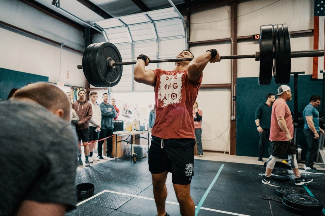 Man lifting a heavy barbell overhead in a CrossFit gym, with a crowd watching in the background.