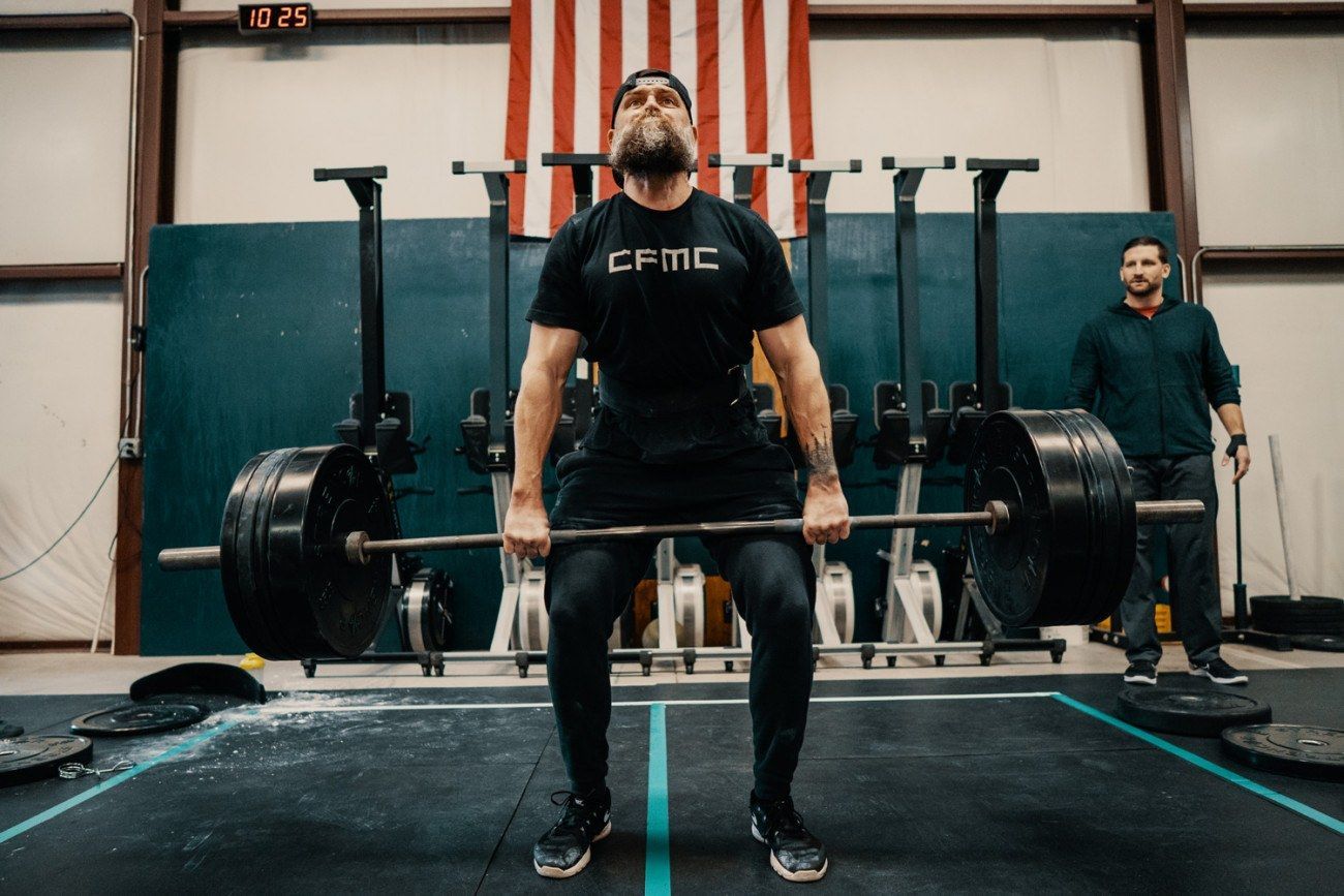Man lifts heavy barbell in a gym, American flag in background, another person watches.