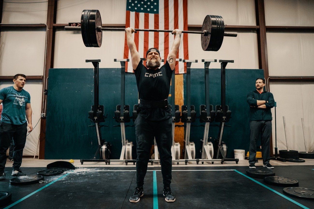 Man overhead presses a barbell in a gym with American flag in the background. Two spotters observe.