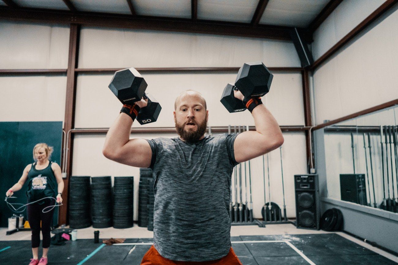 Man lifting dumbbells overhead in a gym. Woman jumps rope in background.