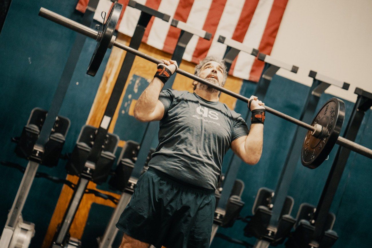 Man lifting barbell overhead in gym, American flag in background.