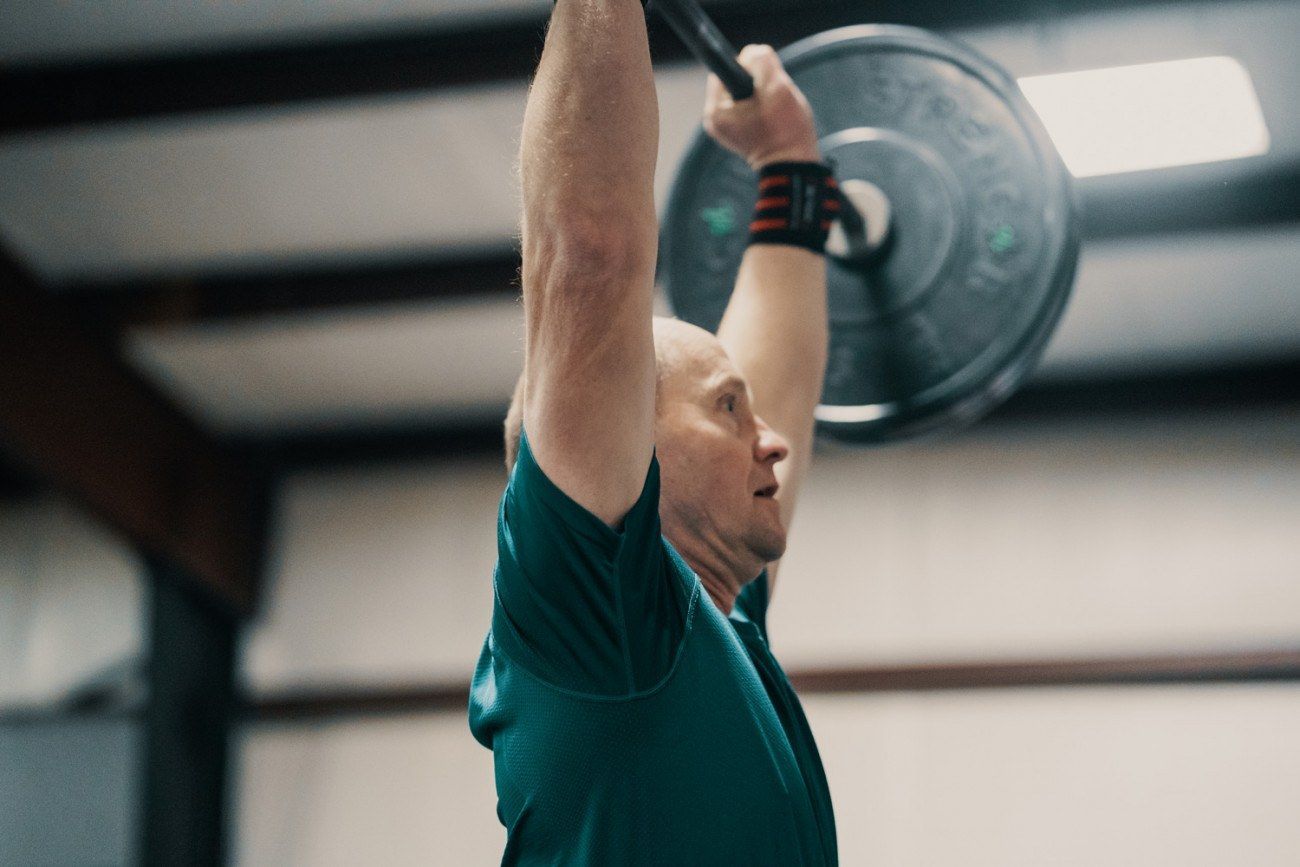 Man lifting barbell overhead in gym. He wears a green shirt and wrist wraps.
