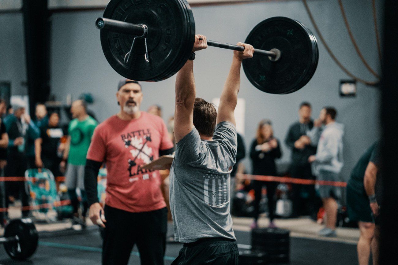 Weightlifter overhead pressing barbell in a gym, coach watches, crowd in background.