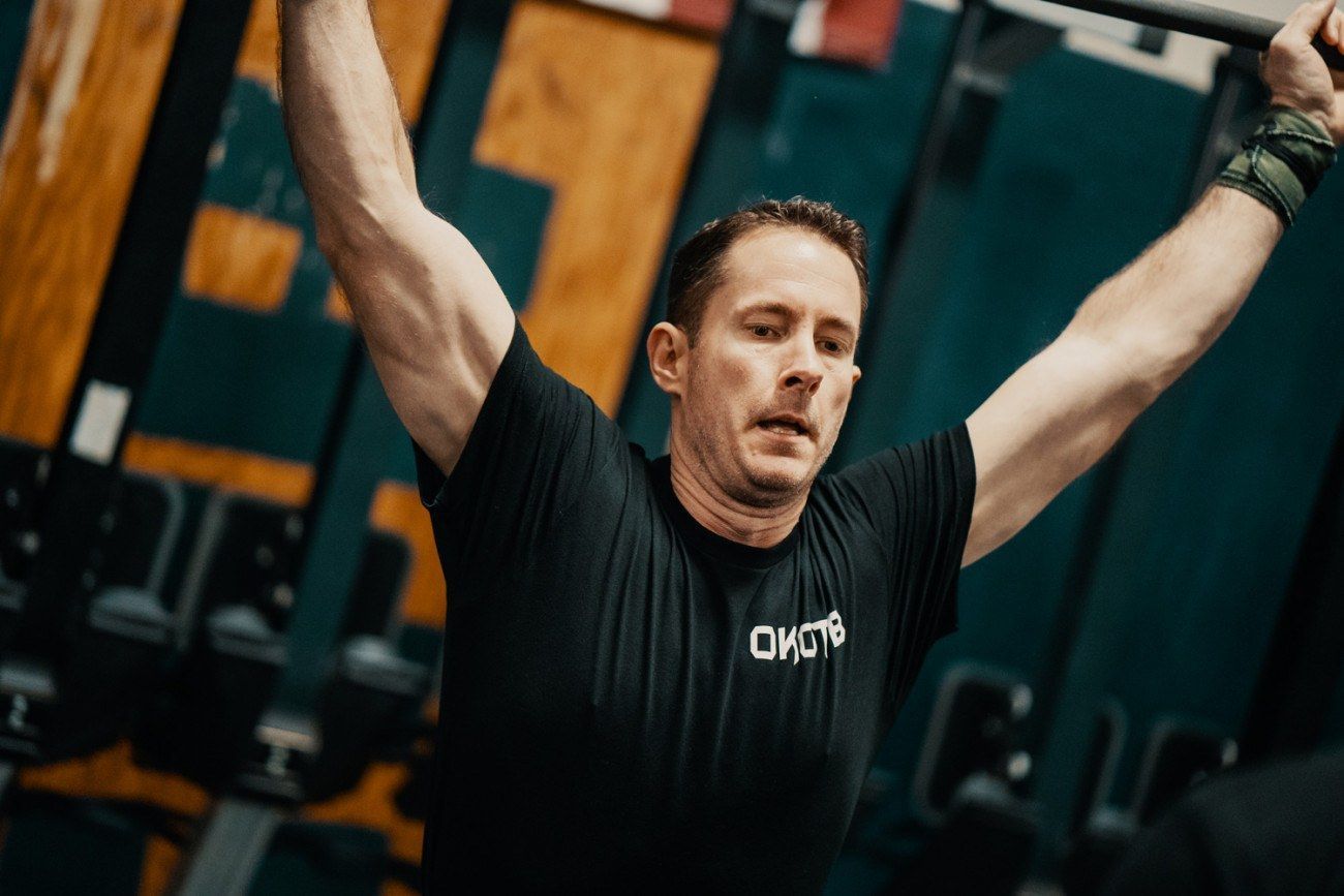 Man in black shirt lifting barbell overhead in gym.