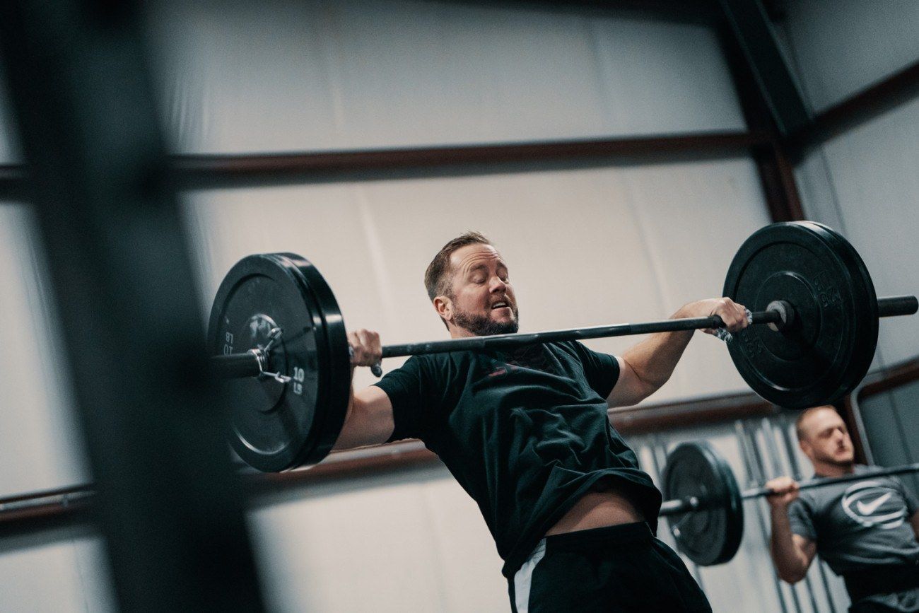 Man lifting barbell overhead in a gym; another man in background.