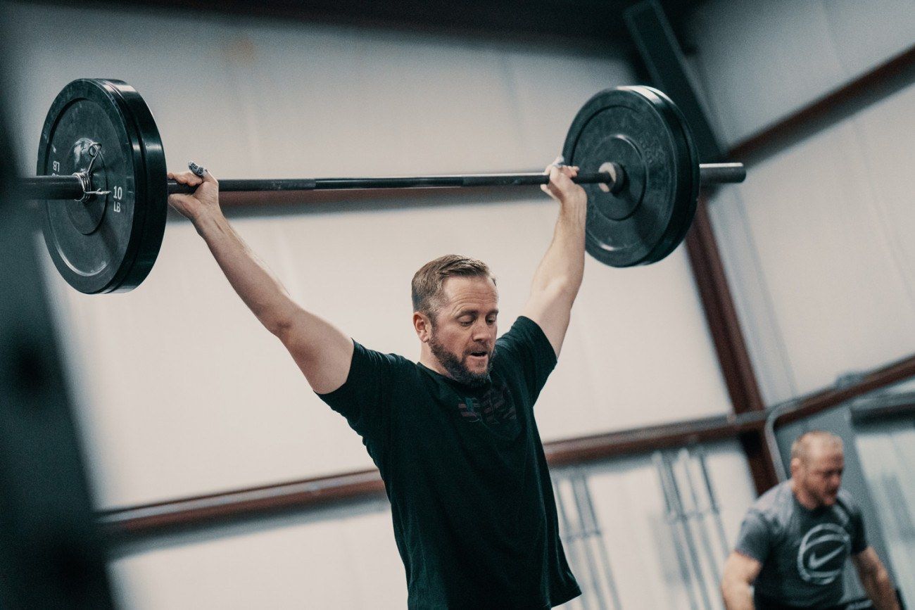 Man lifting a barbell overhead in a gym. Dark shirt, focused expression, weight plates on bar. Another person in background.