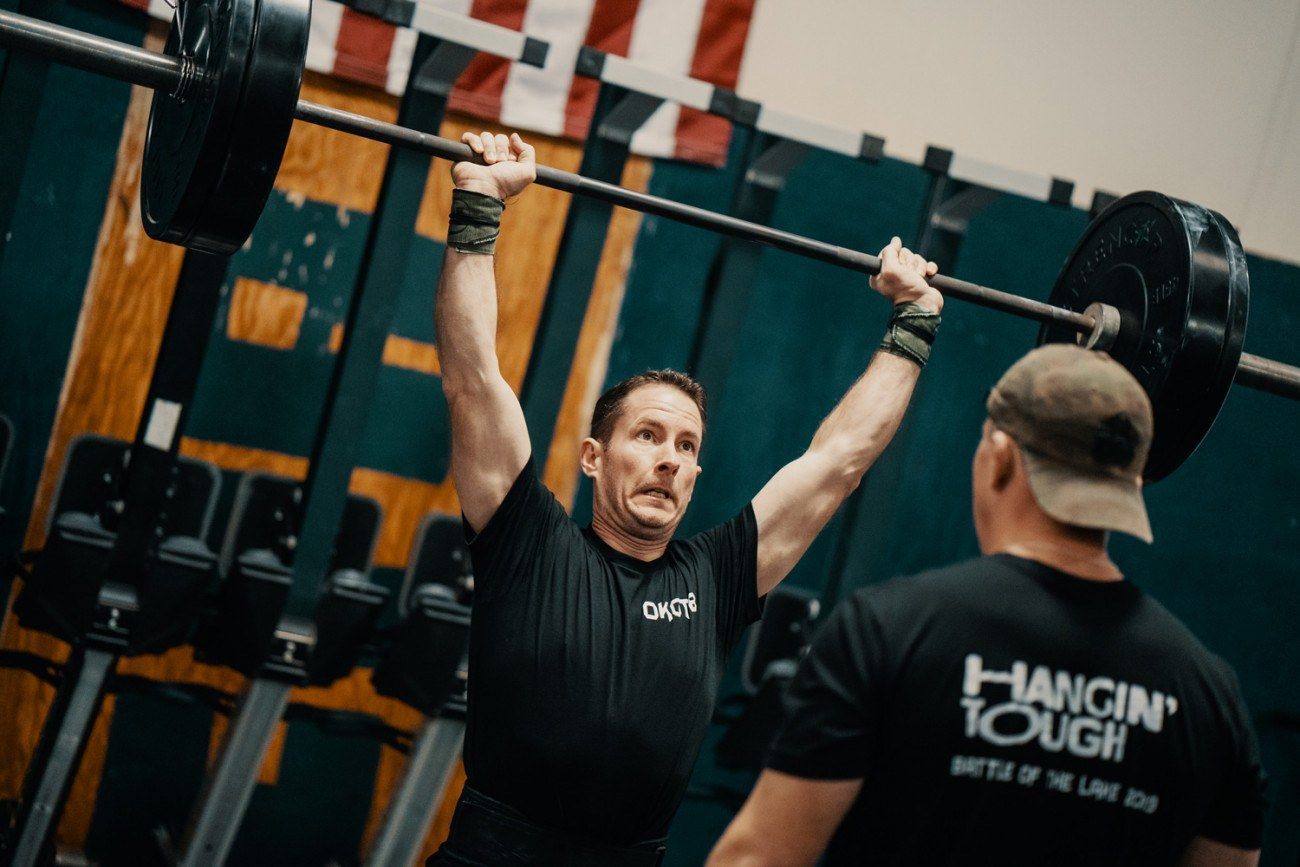 Man lifting barbell overhead with spotter in a gym.