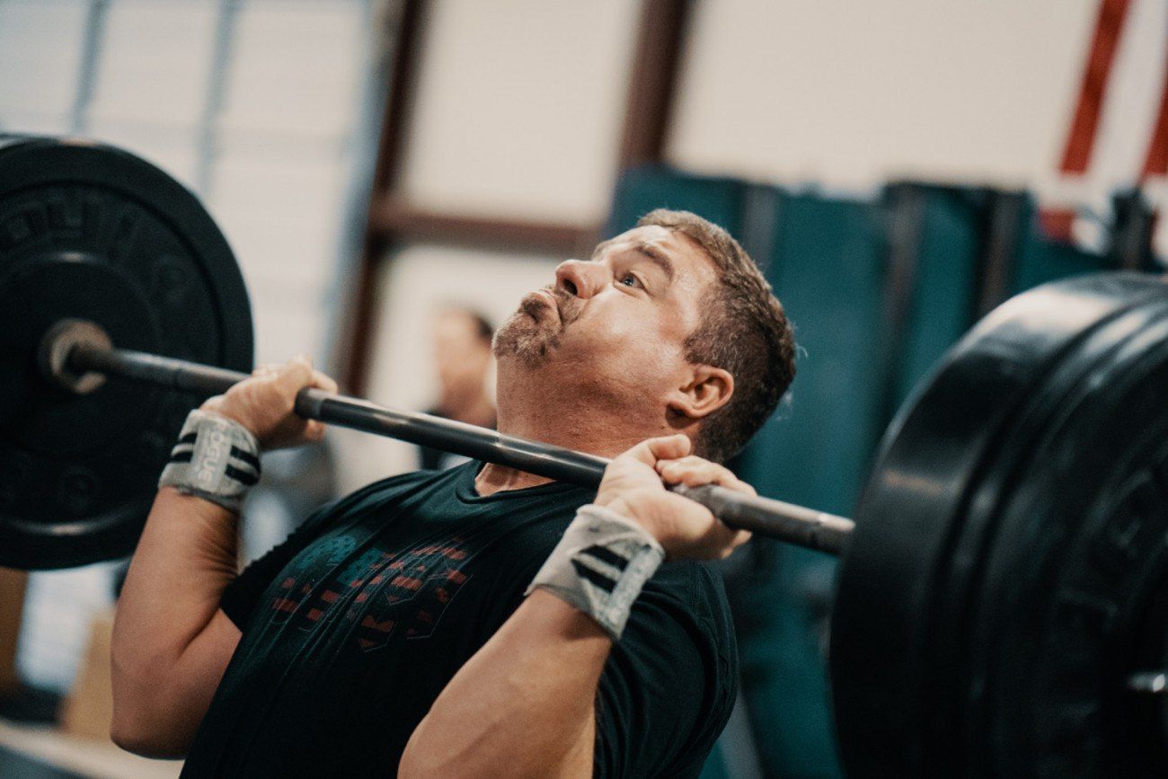 Man straining to lift a barbell overhead, wearing wrist wraps and a black t-shirt, in a gym.
