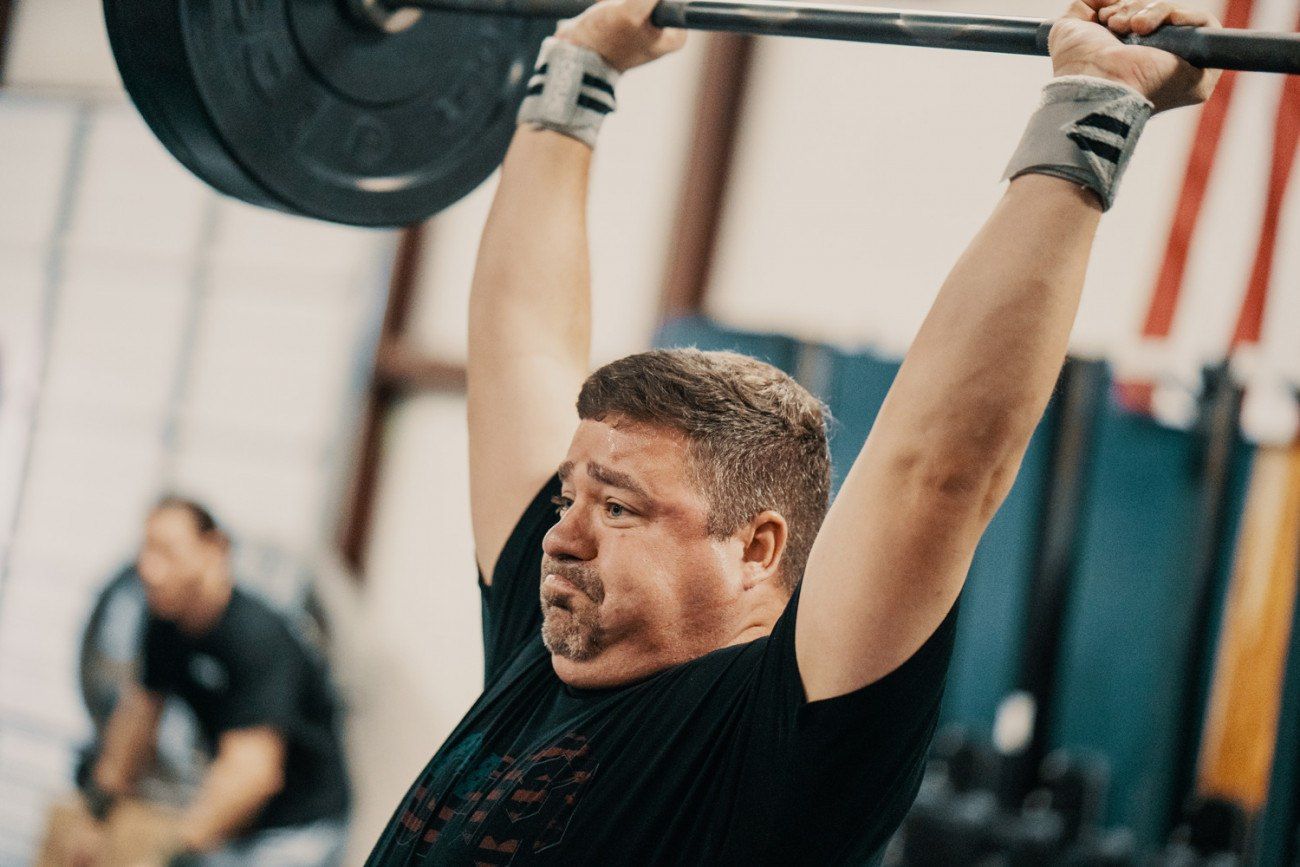 Man lifting barbell overhead in a gym, wearing wrist wraps; focused expression.